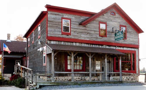 A rustic wooden building with red trim and a sign reading "Jail House," featuring a porch and an American flag nearby.
