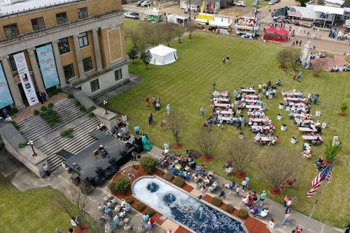 Aerial view of a community event with people seated at tables, a stage, and food vendors in the background.