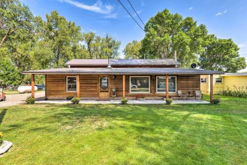 A wooden house with a porch, surrounded by green grass and trees, under a clear blue sky.