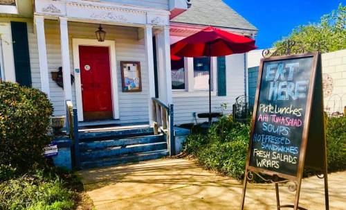 A charming restaurant entrance with a red door, outdoor seating, and a sign listing menu items like sandwiches and salads.