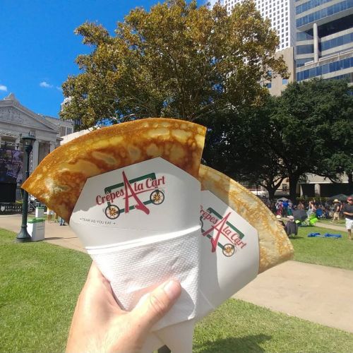A hand holds two crepes in paper wrappers against a sunny park backdrop with trees and buildings.