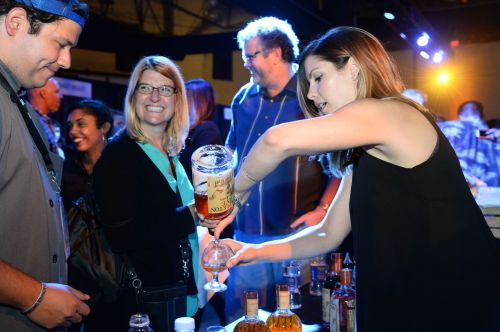 A bartender pours a drink for a smiling guest at a lively event, with people enjoying the atmosphere in the background.