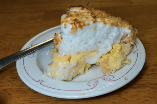 A slice of coconut cream pie topped with toasted coconut on a white plate with a silver fork beside it.