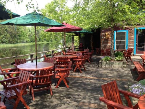 Outdoor seating area with red wooden tables and chairs, shaded by green umbrellas, beside a calm water view.