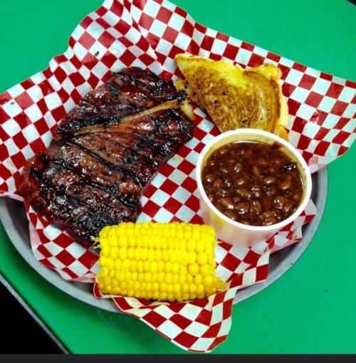 A plate with grilled ribs, baked beans, corn on the cob, and a slice of garlic bread, all on a checkered paper.
