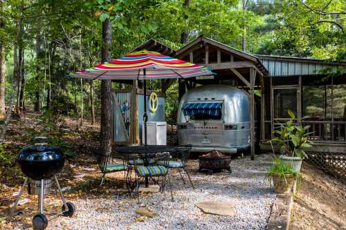 A cozy outdoor space featuring an Airstream trailer, a grill, a table with chairs, and a colorful umbrella in a wooded area.