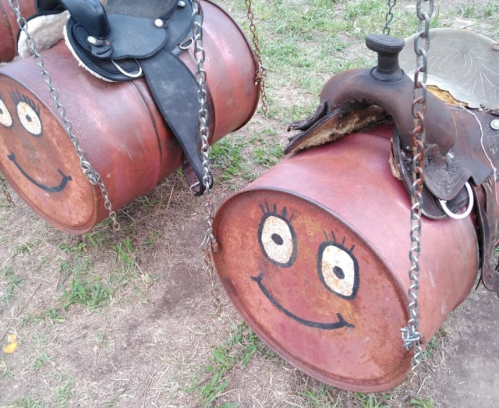 Two smiling barrels with painted faces, hanging from chains, next to saddles on a grassy area.