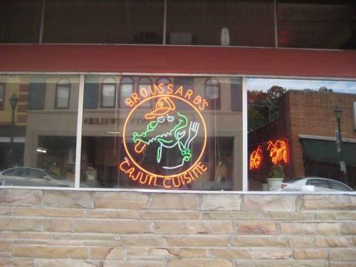 Neon sign for Broussard's Cajun Cuisine featuring an alligator and fork, displayed in a restaurant window.