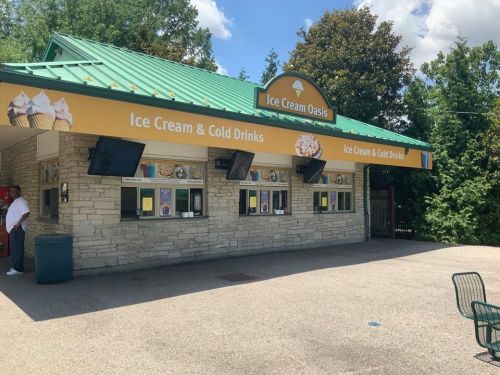 A colorful ice cream stand with a green roof, offering ice cream and cold drinks, surrounded by trees and blue sky.