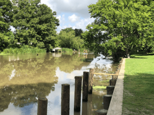 A serene river scene with wooden posts, lush greenery, and people walking along the water's edge under a blue sky.