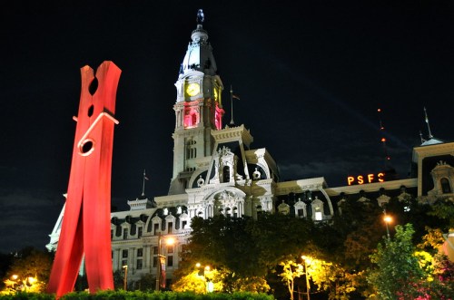 A large red clothespin sculpture in front of a historic clock tower building at night, illuminated by city lights.