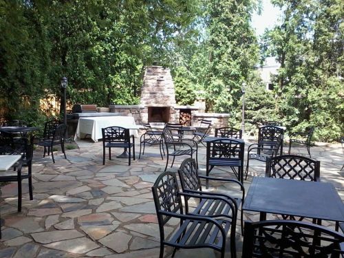 Outdoor patio with stone flooring, black metal tables and chairs, and a stone fireplace surrounded by greenery.