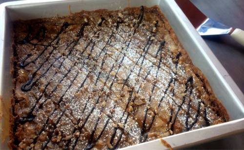 A square pan of dessert topped with powdered sugar and chocolate drizzle, featuring a rich, textured surface.