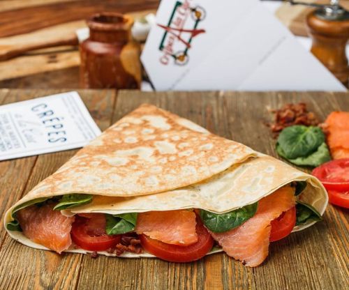 A delicious crepe filled with smoked salmon, spinach, and tomatoes, resting on a wooden table.