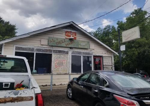 A small, rustic diner with a sign reading "Al's Deli Market" and "Stamps Burger," surrounded by trees and parked cars.