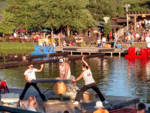 Three performers engage in a lumberjack show on a floating stage by a lake, with an audience in the background.