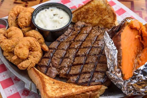 A platter featuring grilled steak, fried shrimp, a baked sweet potato, toast, and a side of creamy coleslaw.