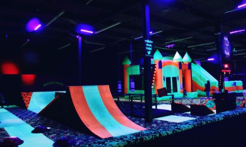 A vibrant indoor trampoline park with colorful inflatable structures and glowing ramps under neon lights.