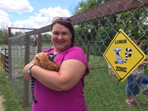 A woman in a pink shirt holds a lemur while standing near a "Lemur Zone" sign in a fenced area.