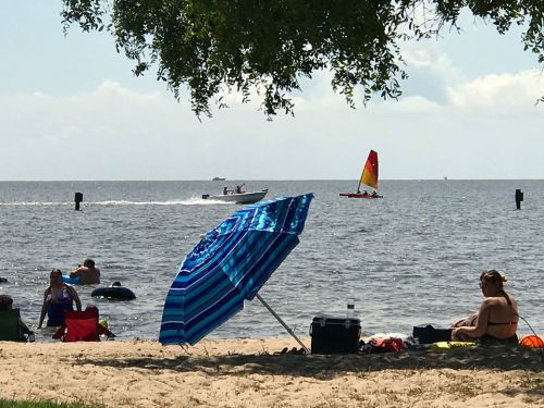 A beach scene with people relaxing, a blue striped umbrella, and a sailboat in the distance on calm waters.