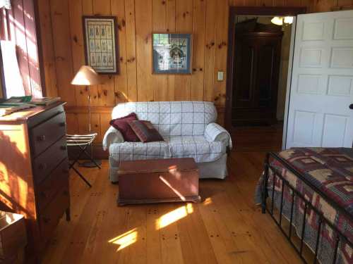 Cozy wooden bedroom with a white couch, quilted bed, dresser, and warm sunlight streaming through the window.