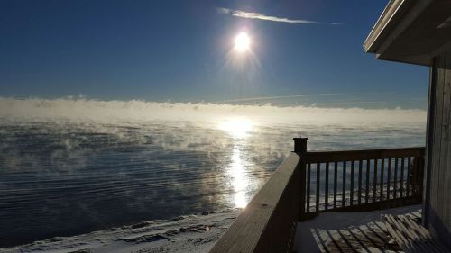 A serene winter scene of a sunlit lake with mist rising, viewed from a wooden deck.
