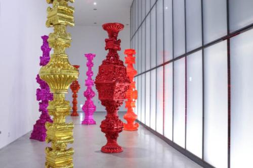 Colorful, large sculptures in vibrant pink, gold, and red arranged in a modern gallery space with glass walls.