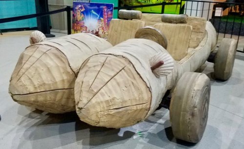 A whimsical car sculpture made of wood, featuring rounded, log-like shapes for wheels and a playful design.