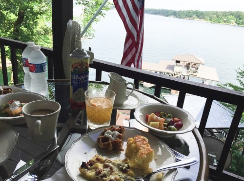 A lakeside breakfast table with coffee, fruit, waffles, and a view of the water and trees, featuring an American flag.