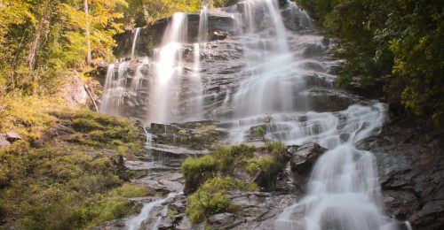 A cascading waterfall flows over rocky terrain, surrounded by lush greenery and autumn-colored trees.