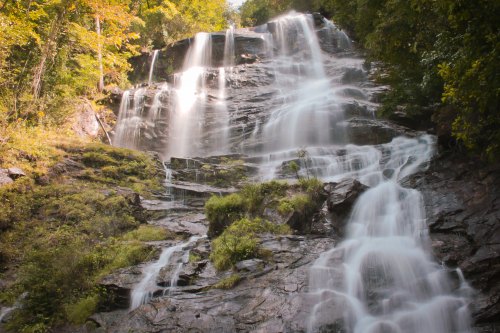 A cascading waterfall flows over rocky terrain, surrounded by lush greenery and autumn-colored trees.