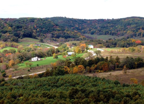 A scenic view of rolling hills with colorful autumn foliage, farms, and a winding road in a rural landscape.