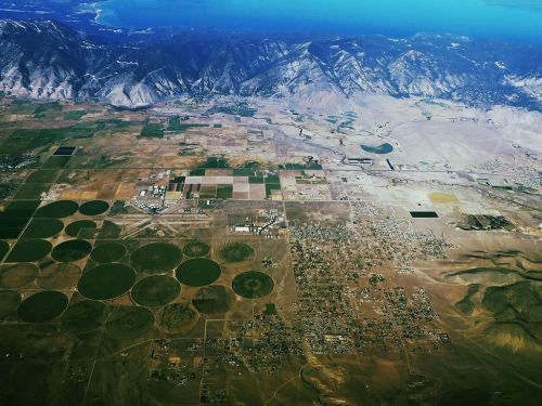 Aerial view of diverse agricultural fields, circular plots, and mountainous terrain near a body of water.