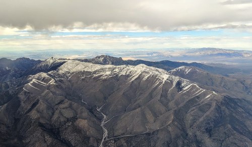 Aerial view of snow-capped mountains under a cloudy sky, with valleys and distant ranges visible in the background.