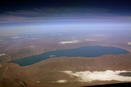 Aerial view of a large lake surrounded by mountains and snow-capped peaks under a clear blue sky.