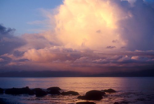 A serene seascape at sunset, featuring calm waters, rocky shorelines, and dramatic clouds illuminated by warm light.