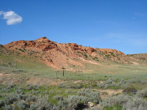 A rocky hillside with reddish formations under a clear blue sky, surrounded by green grass and sparse vegetation.