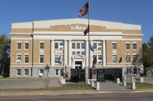 A large brick courthouse with columns, flags, and a statue in front, set against a clear blue sky.