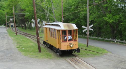 A vintage yellow streetcar on tracks near a crossing, surrounded by greenery and a gravel path.