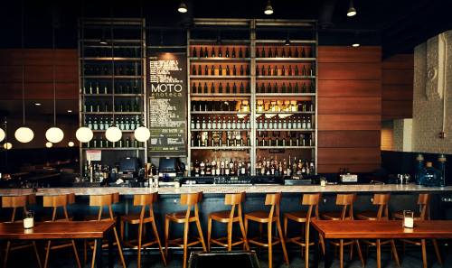 A modern bar with wooden stools, a large shelf of bottles, and a menu board in a stylish restaurant setting.
