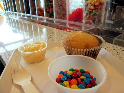 A tray with a muffin, a cup of yellow frosting, and a cup of colorful candy-coated chocolates.