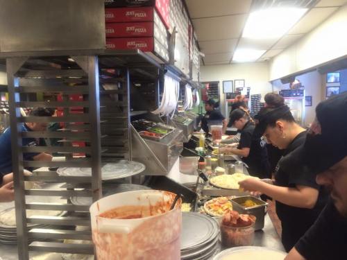 A busy kitchen with staff preparing food, including pizzas and salads, surrounded by various ingredients and utensils.