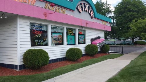 A colorful pizza and burger restaurant with neon signs, featuring a drive-thru and well-maintained landscaping.