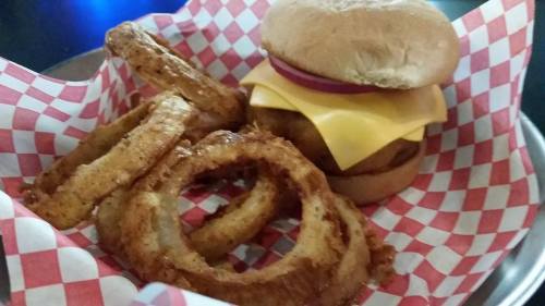 A basket with a cheeseburger topped with cheese and onion, alongside crispy onion rings on a checkered paper liner.