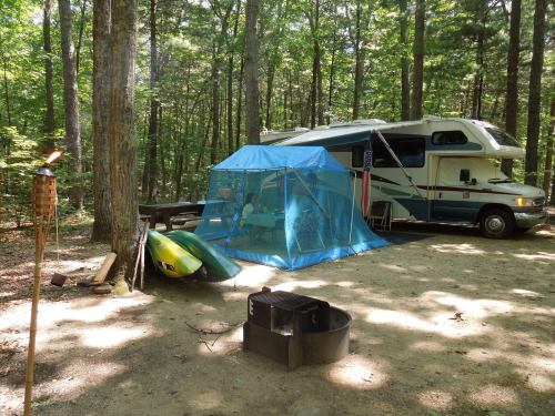 A camper parked in a wooded area, with a blue tent, kayaks, and a fire pit nearby.