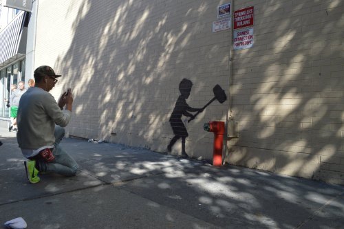 A person photographs a shadowy mural of a child with a hammer next to a fire hydrant on a city street.