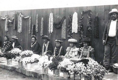 A group of people in traditional attire, adorned with floral garlands, seated in front of a wooden wall decorated with flowers.