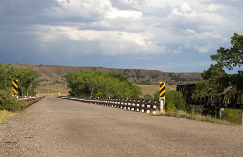 A gravel road leads to a wooden bridge, surrounded by green trees and rolling hills under a cloudy sky.