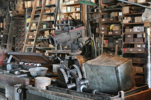 A cluttered workshop filled with various tools, machinery, and storage boxes on shelves in the background.
