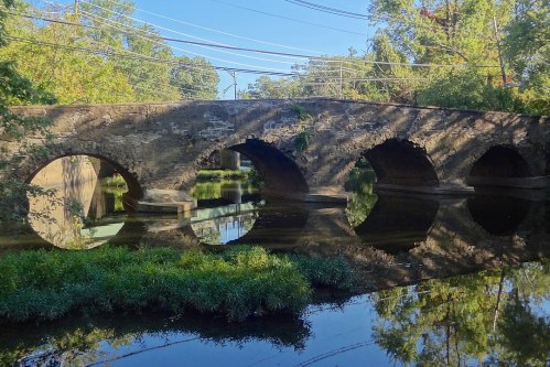 A stone bridge with arches spans a calm river, surrounded by lush greenery and blue skies. Reflections are visible in the water.